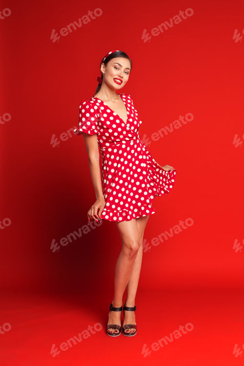 Preview: Model poses in red polka dot dress against bright red background in stylish studio setting