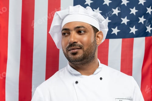 Preview: Young professional chef in white cap and coat standing against American flag