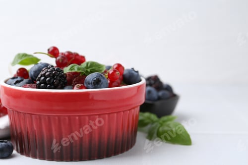 Preview: Different ripe berries and basil leaves in bowl on white tiled table, closeup. Space for text