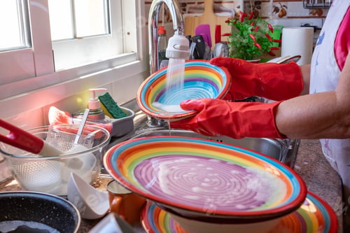 Preview: Elderly woman washing crockery in front to the window, wearing red gloves. Home kitchen