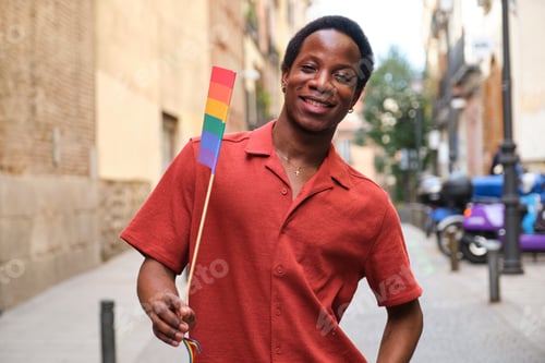 Preview: Gay black man in a red shirt is holding a rainbow flag and smiling at Pride Day.