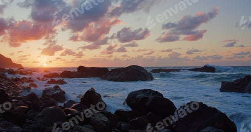 Preview: Sunset over ocean. Orange clouds and silhouette of rock formations, mountains. Rocky beach, waves