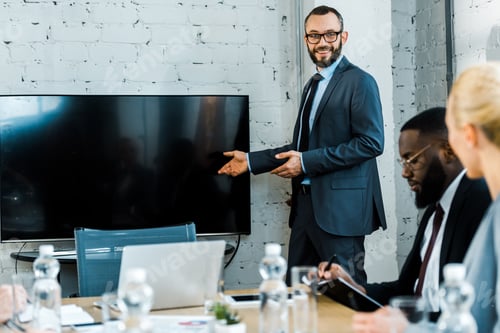 Preview: cheerful business coach in eye glasses gesturing near tv with blank screen and multicultural