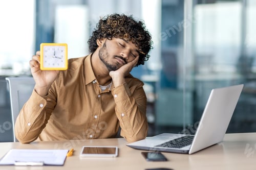 Preview: Exhausted muslim man sitting at laptop with closed eyes and showing alarm clock in one hand. Young