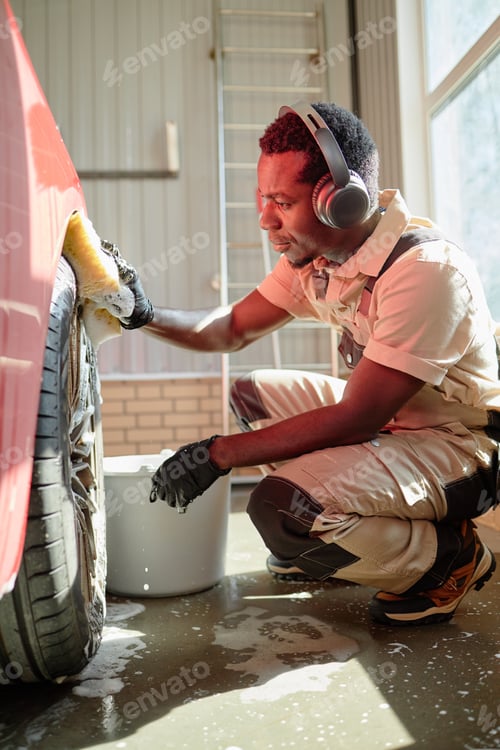 Preview: Young Adult Black Man Wearing Headphones Washing Car Tire Indoors