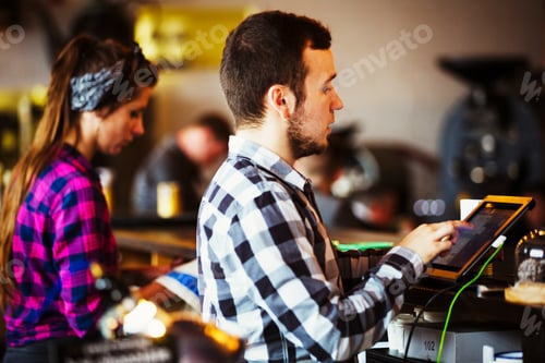 Preview: Specialist coffee shop. A woman and man working behind the counter by a roasting machine.