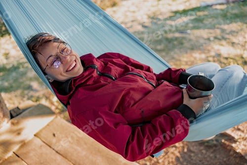 Preview: Portrait of young cheerful happy woman in glasses lying in hammock, smiling and holding cup.