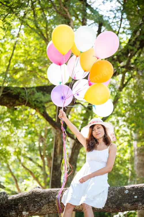 Preview: Cheerful beauty woman holding balloons relax sitting under big tree in green park with happiness