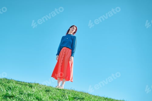 Preview: Teen girl in blue top and coral skirt standing on a grassy hill against a blue sky.