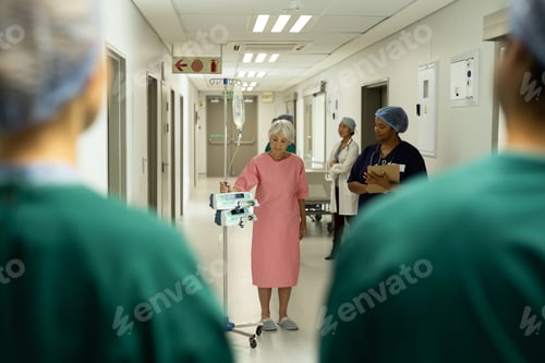 Preview: Senior caucasian female patient with iv pole on wheels walking in busy hospital corridor