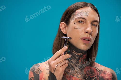 Preview: tattooed young man with piercing holding safety razor and shaving face isolated on blue