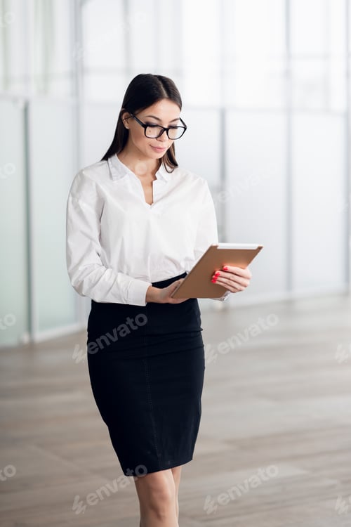 Preview: Attractive young woman wearing glasses and reading her touchscreen tablet while standing inside