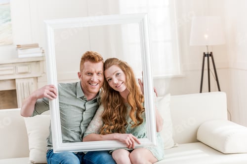 Preview: Beautiful young couple sitting embracing on sofa and looking through white frame