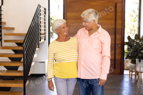 Aperçu: Smiling biracial senior friends looking at each other while standing against door in nursing home