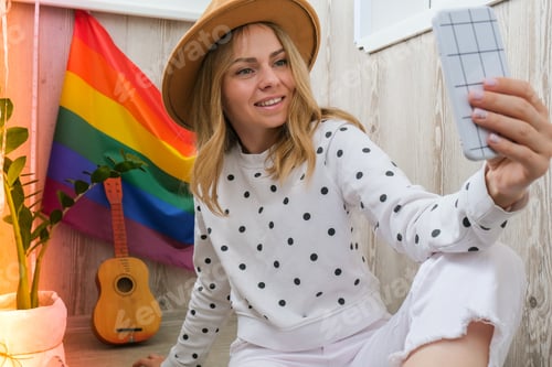 Preview: Young millennial hippie woman sitting on balcony using mobile phone. LGBTQ rainbow flag on backgroun