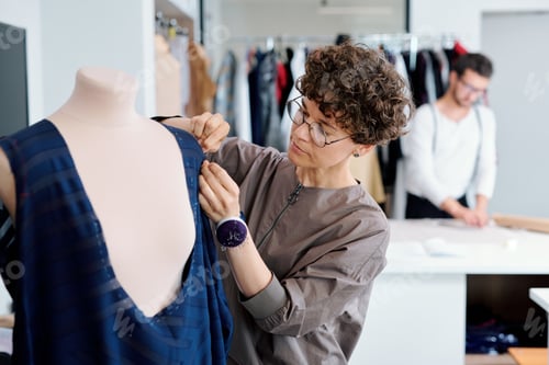 Preview: Young serious seamstress stitching unfinished blue dress on mannequin