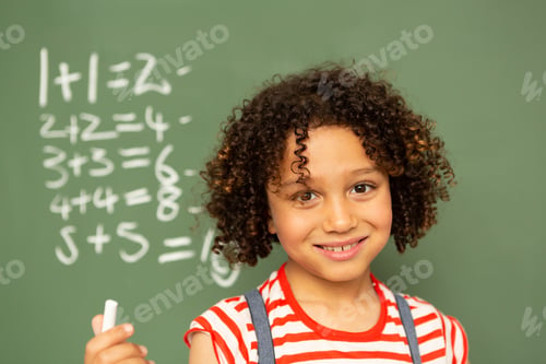 Preview: Mixed-race schoolgirl standing against green board and holding a chalk in a classroom