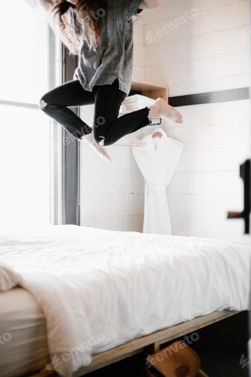 Preview: Young woman with long hair and casual clothing jumping on a bed in a modern and minimal hotel room