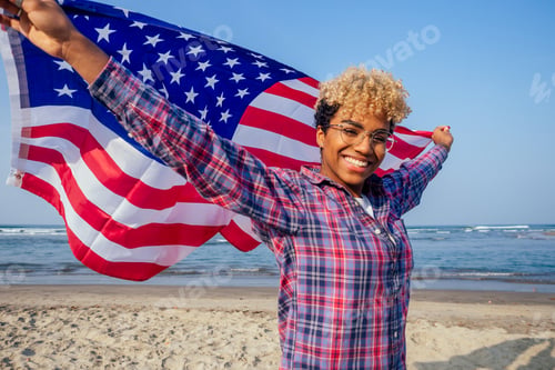 Preview: happy afro american woman holding usa flag at sunny tropical beach.
