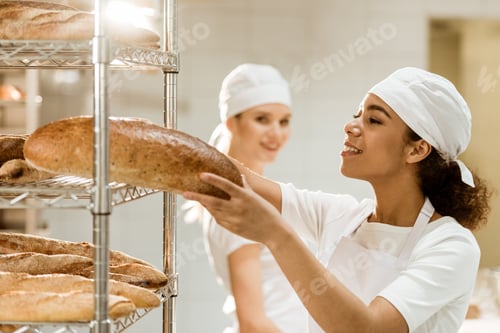 Preview: female baker putting fresh bread loaves on shelves at baking manufacture while her colleague