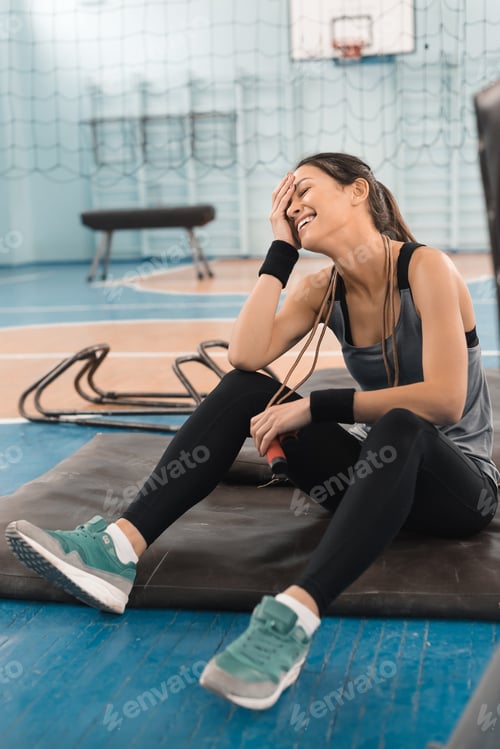 Preview: Attractive young sportswoman sitting on mat and laughing in sports hall