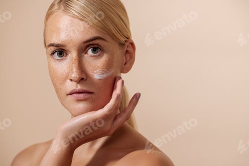 Preview: Young beautiful woman with facial cream on a cheek touching face and looking at camera