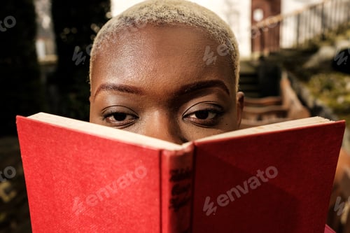 Preview: Young student hiding face behind red book looking at camera