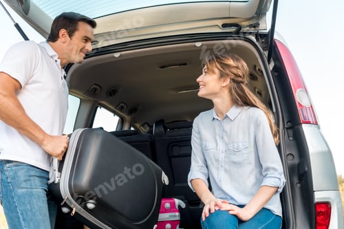 Preview: happy adult man packing luggage into car trunk while his girlfriend looking at him