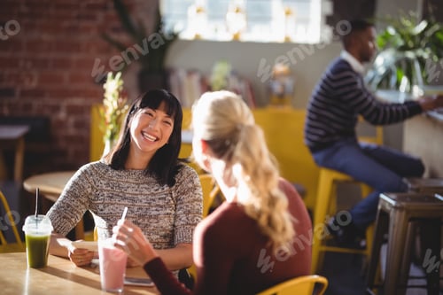 Preview: Cheerful female friends talking while sitting with fresh drinks at table