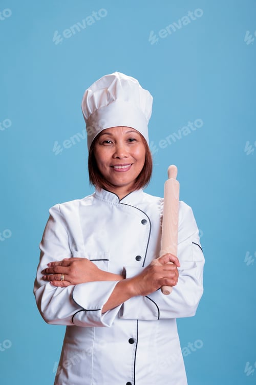 Preview: Professional positive asian cook standing with arm crossed holding kitchen rolling pin