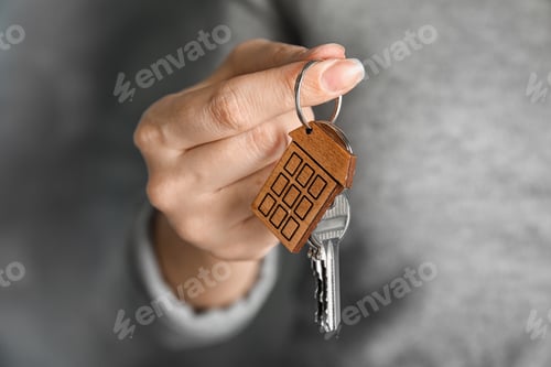 Preview: Woman holding house key with trinket on blurred background, closeup