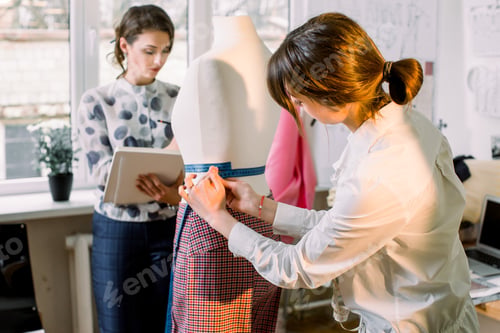 Preview: Back view of female dressmaker measuring mannequin with tape in atelier office.