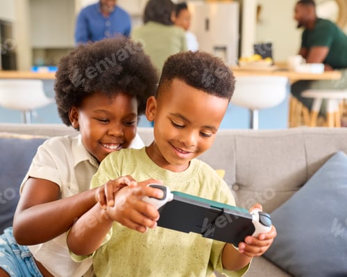 Preview: Boy And Girl Playing With Handheld Gaming Device At Home With Multi Generation Family In Background