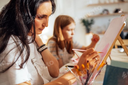 Preview: A young focused woman in a white T-shirt and apron paints a picture at a table and an easel.