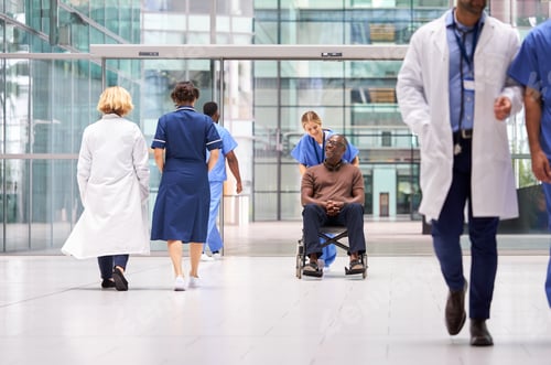 Preview: Female Nurse Wearing Scrubs Wheeling Patient In Wheelchair Through Lobby Of Modern Hospital Building