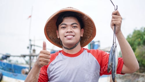 Preview: Happy young fisherman on the beach holding his catch fish and shows in front of his boat