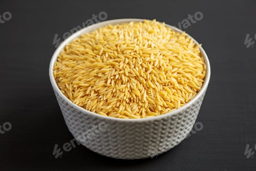 Preview: Raw Organic Risoni Pasta in a Bowl on a black background, side view.