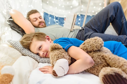 Preview: Smiling father reading book to cute little boy lying and hugging teddy bear