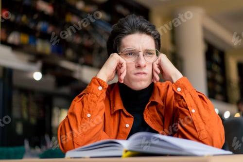 Preview: tired boy student sitting in front of a book in the library bored and lazy while preparing for exams