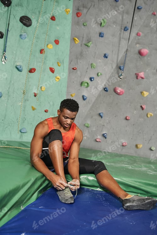 Preview: vertical shot of sporty african american man on crash pad tying his shoes with rock wall on backdrop