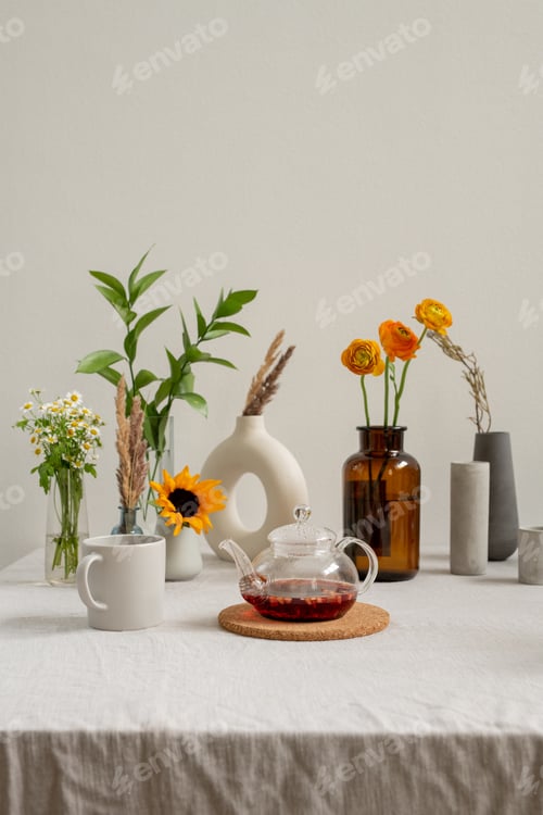 Preview: Teapot with fresh herbal tea and mug on background of various flowers in vases