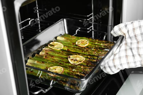 Preview: Person taking glass baking dish with cooked asparagus and lemon slices from oven, closeup