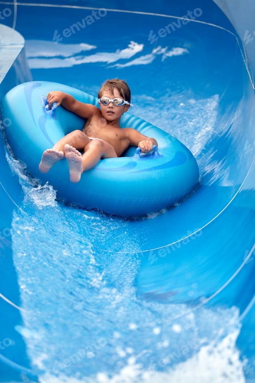 Preview: Joyful boy in swimming goggles slides down the blue water slide on an inflatable boat in the water