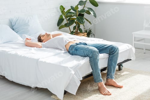 Preview: handsome man in t-shirt and jeans lying on back on bed in bedroom