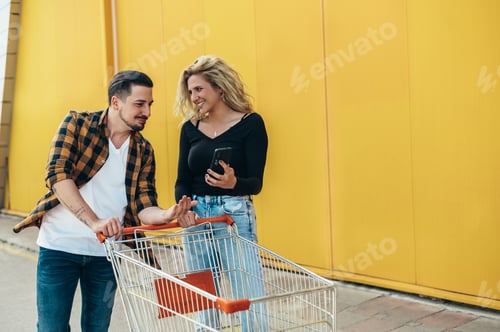 Preview: Couple using smartphone and taking a selfie while holding a supermarket shopping cart