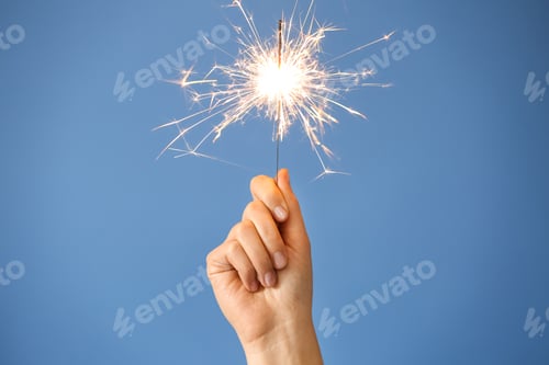 Preview: Woman holding bright burning sparkler on light blue background, closeup