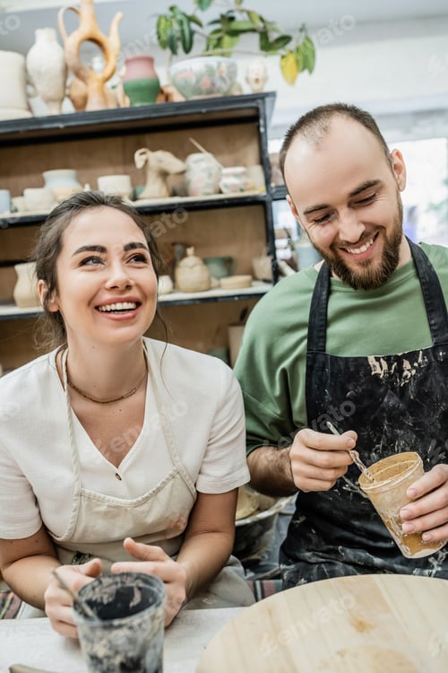 Preview: Cheerful couple of artisans holding dye while working together in ceramic workshop