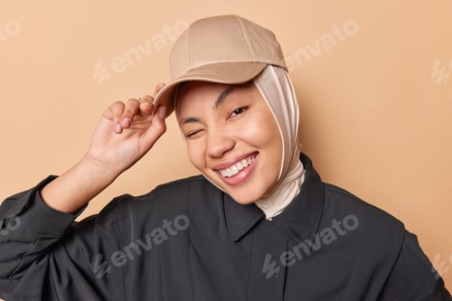Preview: Portrait of happy positive young woman winks eye smiles toothily feels very glad keeps hand on cap
