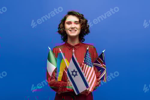 Preview: pleased woman smiling at camera while holding flags of different countries isolated on blue