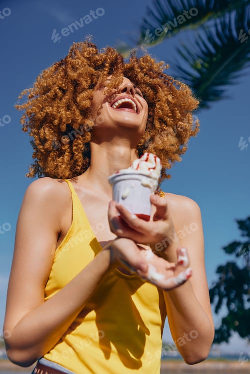 Preview: Joyful woman with curly hair enjoying ice cream outdoors near palm trees